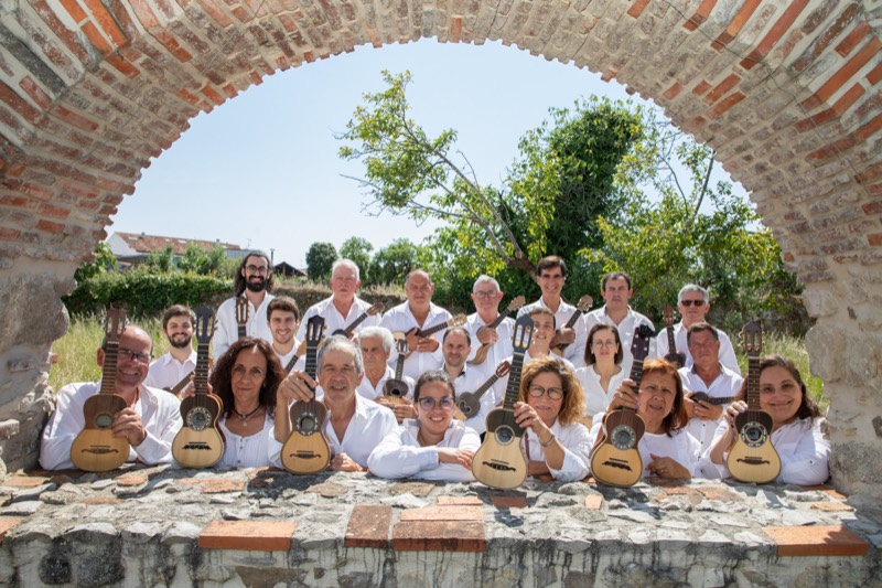 O Grupo de Cavaquinhos do Louriçal debaixo de um arco de pedra e tijolo: cerca de vinte pessoas de camisa branca, apoiadas num muro baixo, cada uma com o seu cavaquinho na mão. Ao fundo, através do arco, vê-se uma árvore e o céu limpo.