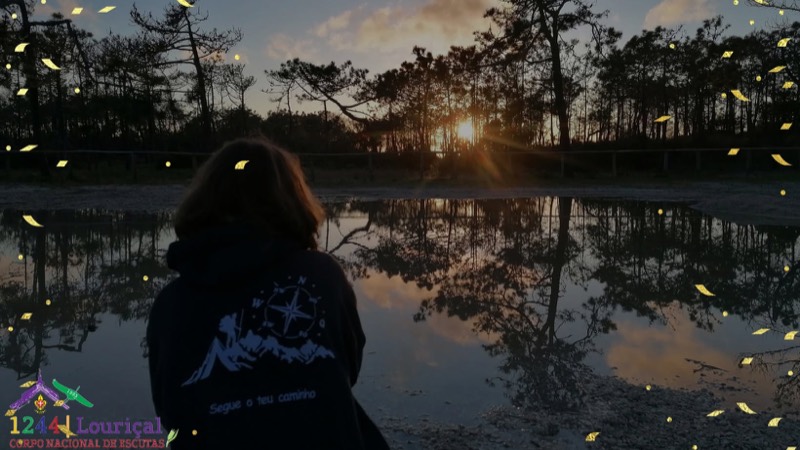 Uma pessoa de costas, de camisola escura com uma rosa dos ventos e montanhas estampadas por cima da frase «Segue o teu caminho», olha uma lagoa ao pôr do sol que reflete os pinheiros da margem. No canto inferior esquerdo, aplicado sobre a fotografia, o emblema do Agrupamento 1244 do Louriçal.