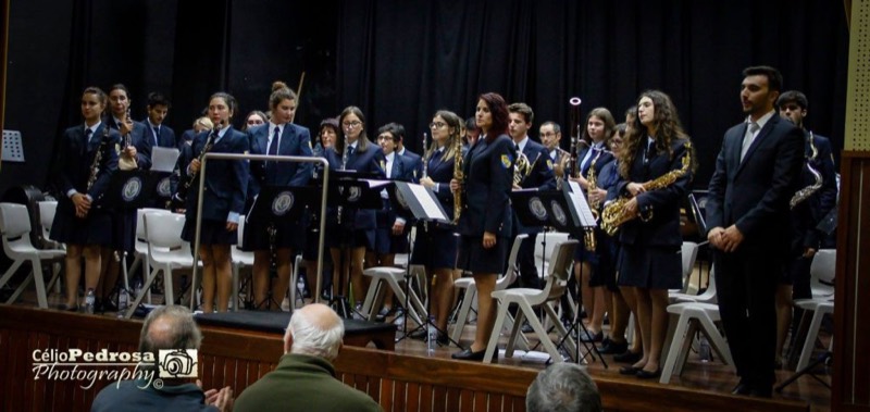 A Sociedade Filarmónica Louriçalense num palco: cerca de trinta músicos de fato azul-escuro, de pé atrás das estantes de partituras, com clarinetes, flautas e saxofones nas mãos. Ao fundo, uma cortina preta; em primeiro plano, as cabeças das pessoas na plateia.