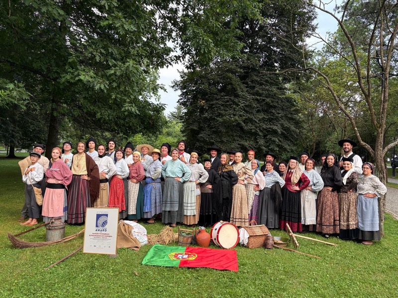 O Rancho Folclórico e Etnográfico do Louriçal num relvado, à sombra de árvores altas: cerca de quarenta pessoas em traje tradicional, com saias de chita, aventais, xailes, lenços na cabeça e chapéus pretos. Pousados na relva à frente, a bandeira portuguesa, um bombo, um cântaro de barro, cestas de verga, ancinhos e um diploma emoldurado.
