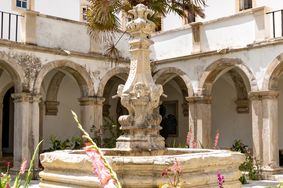 O claustro do Convento: um pátio rodeado por arcarias de pedra em dois pisos, com um chafariz de pedra lavrada ao centro, entre canteiros de flores e uma palmeira.