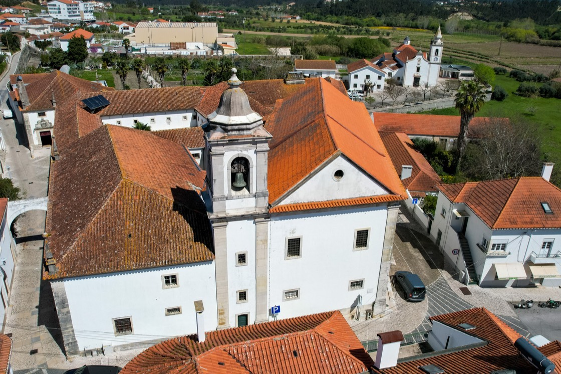 O Convento visto do ar: o corpo da igreja ao centro, com a torre sineira e o sino à vista, rodeado pelos telhados de telha do resto do edifício. À volta, as casas do Louriçal e os campos, e ao fundo outra igreja de fachada branca.