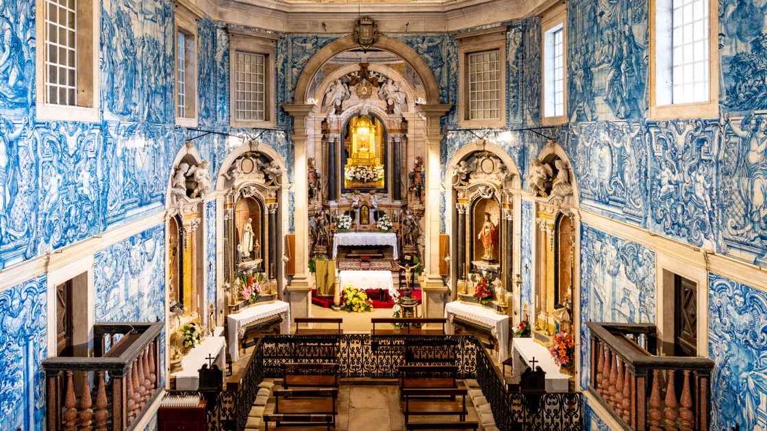 O interior da igreja do Convento, visto de cima: as paredes cobertas de azulejo azul e branco do chão ao teto, o altar-mor em talha dourada ao fundo com uma imagem num nicho iluminado e flores à sua frente, dois altares laterais e, em primeiro plano, a grade de ferro e os bancos de madeira.