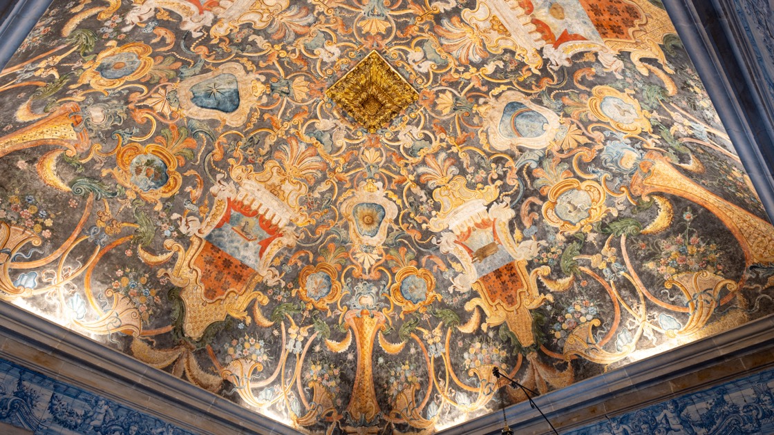 O teto pintado da igreja, visto de baixo: uma composição simétrica de volutas, conchas, flores e medalhões em ocre, azul e vermelho sobre fundo escuro, com um elemento dourado em relevo ao centro. Em baixo, o topo dos painéis de azulejo.