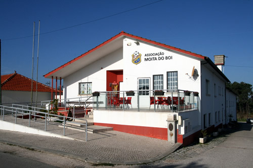 A sede da Associação da Moita do Boi: um edifício branco de dois andares com faixas e beirados vermelhos, o nome e o emblema da associação na fachada e, no andar de cima, uma esplanada coberta com mesas e cadeiras vermelhas atrás de uma guarda de vidro. À frente, a rampa e as escadas de acesso, e dois mastros de bandeira.