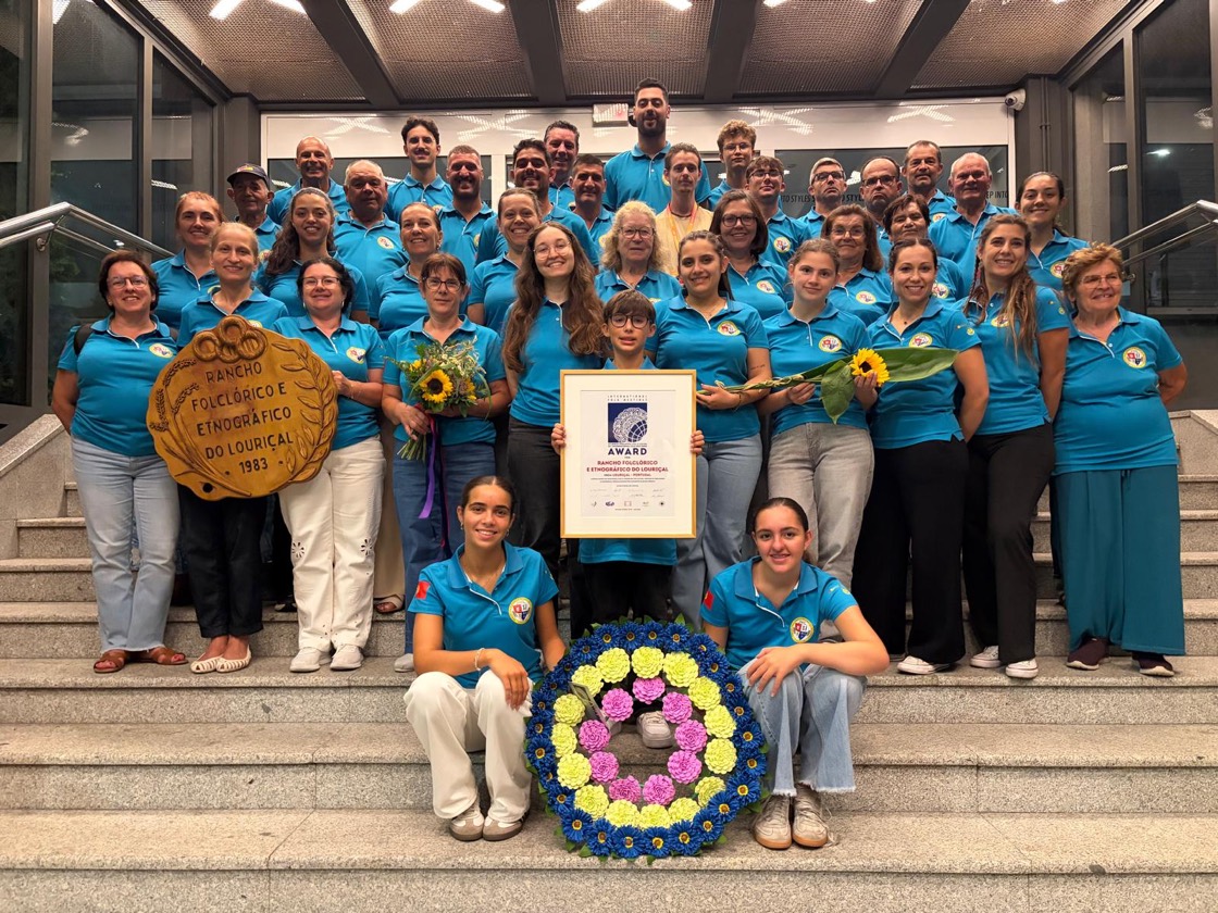 Fotografia de grupo do Rancho Folclórico e Etnográfico do Louriçal numa escadaria: cerca de quarenta pessoas, de todas as idades, com o mesmo pólo azul-claro e o emblema do rancho ao peito. À frente seguram uma placa de madeira esculpida com o nome do rancho e a data de 1983, um diploma emoldurado, ramos de girassóis e uma coroa de flores azuis, cor-de-rosa e amarelas.
